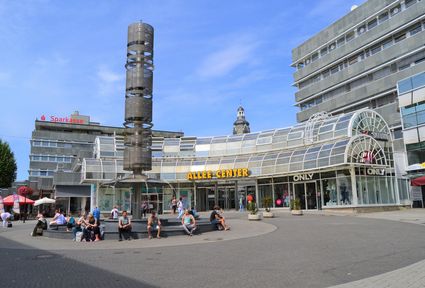 Haupteingang des Einkaufscenters Allee Centers mit einem Brunnen im Vordergrund, auf dessen Rand mehrere Menschen sitzen.